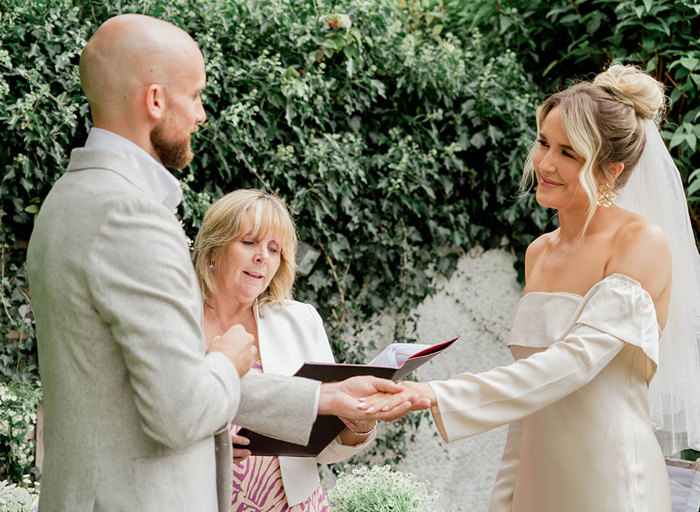 a bride and groom look lovingly at one another as groom makes ring exchange. A celebrant stands between them and there is greenery in background