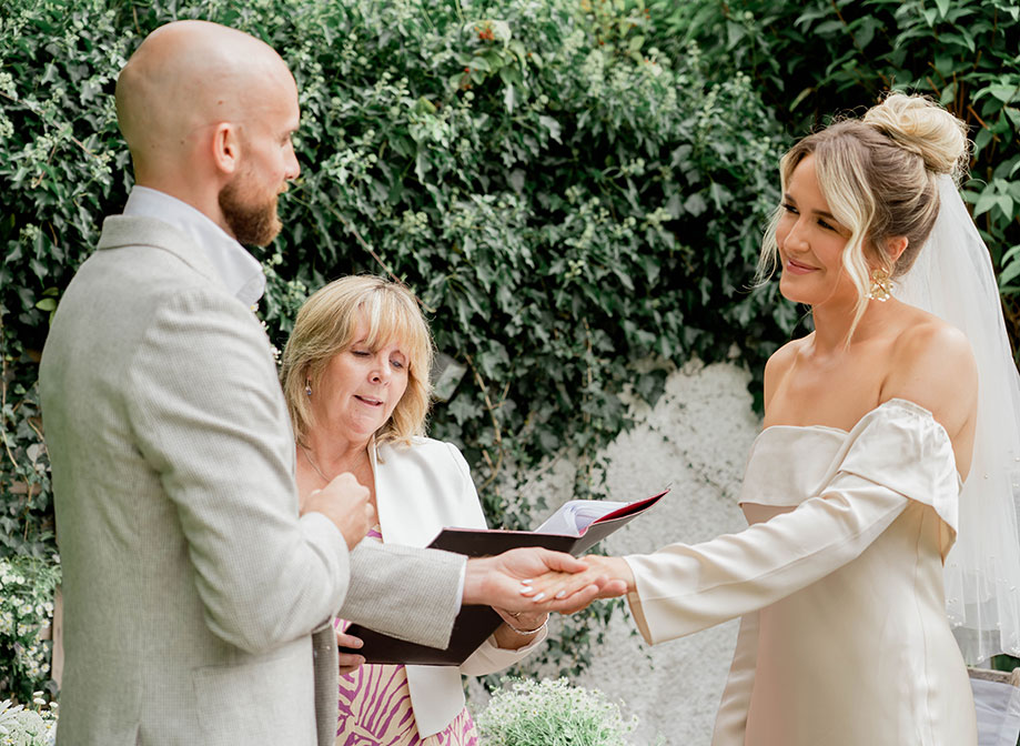 a bride and groom look lovingly at one another as groom makes ring exchange. A celebrant stands between them and there is greenery in background