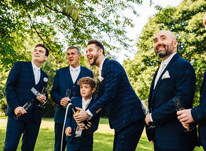 Groomsmen lined up in matching suits pop champagne with the groom and his young son