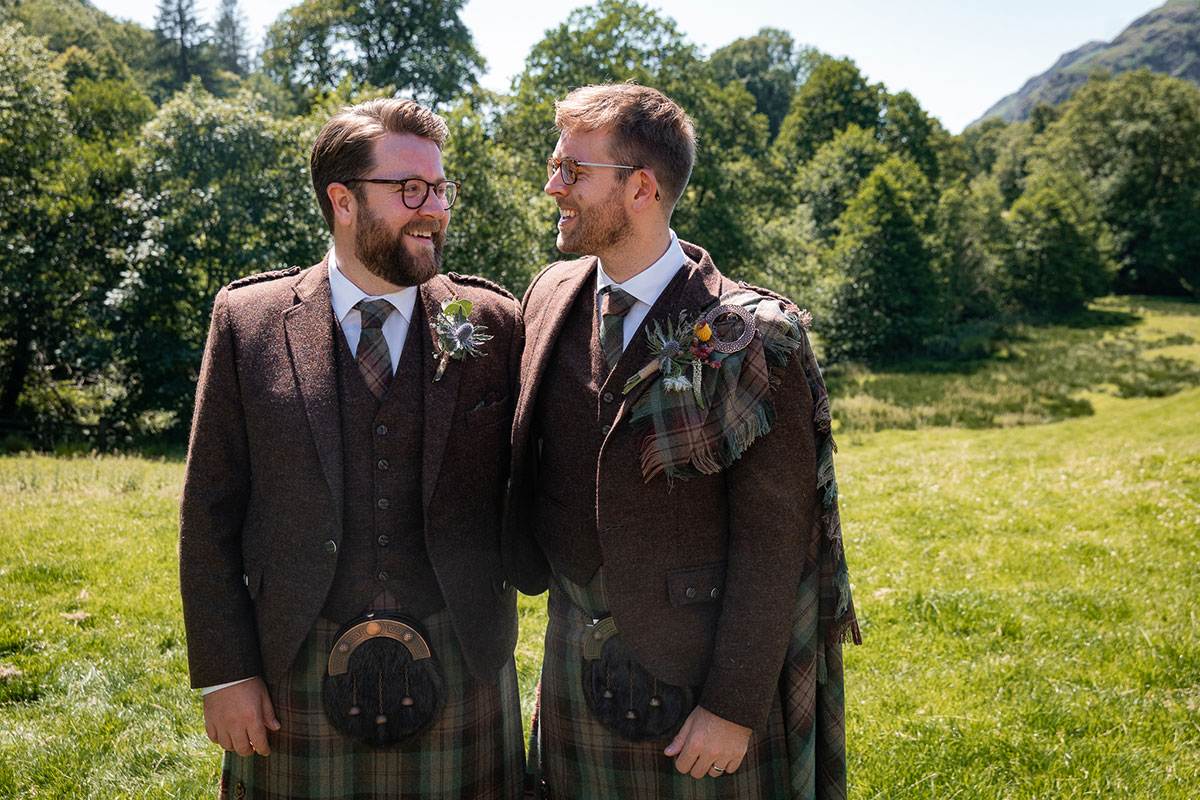 Groom and best man stand side by side outdoors in a green countryside setting, wearing matching brown tweed jackets with tartan kilts and sporrans