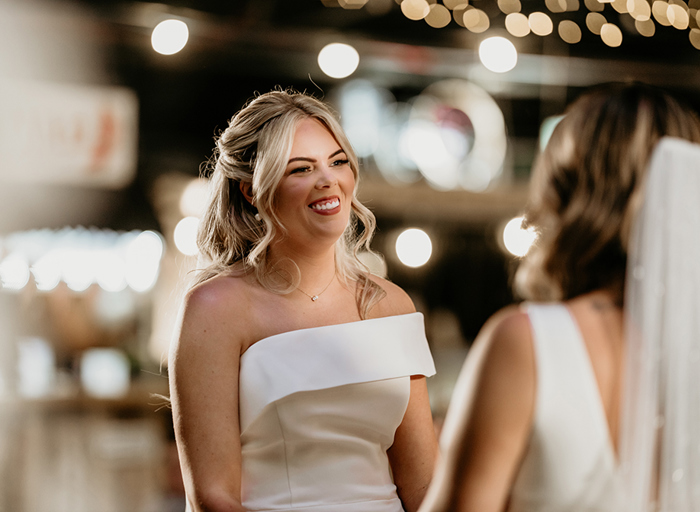 a smiling bride looks at another bride with blurred out lights in the background