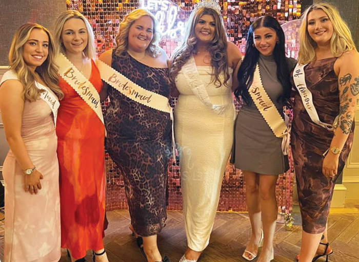 Five women all in long dresses and sashes on lit background