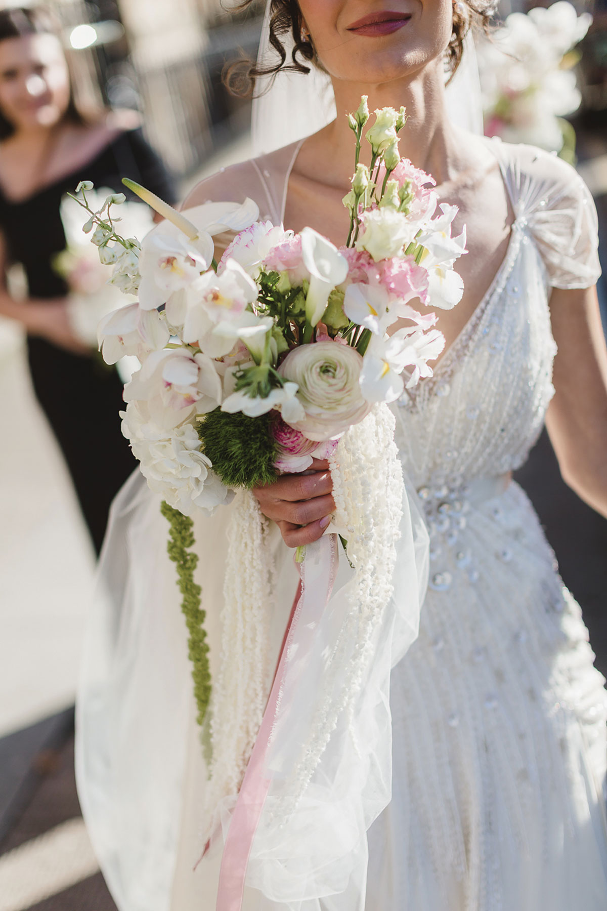 Bride in beaded gown holding a soft white and blush wedding bouquet featuring orchids, calla lilies and trailing amaranthus