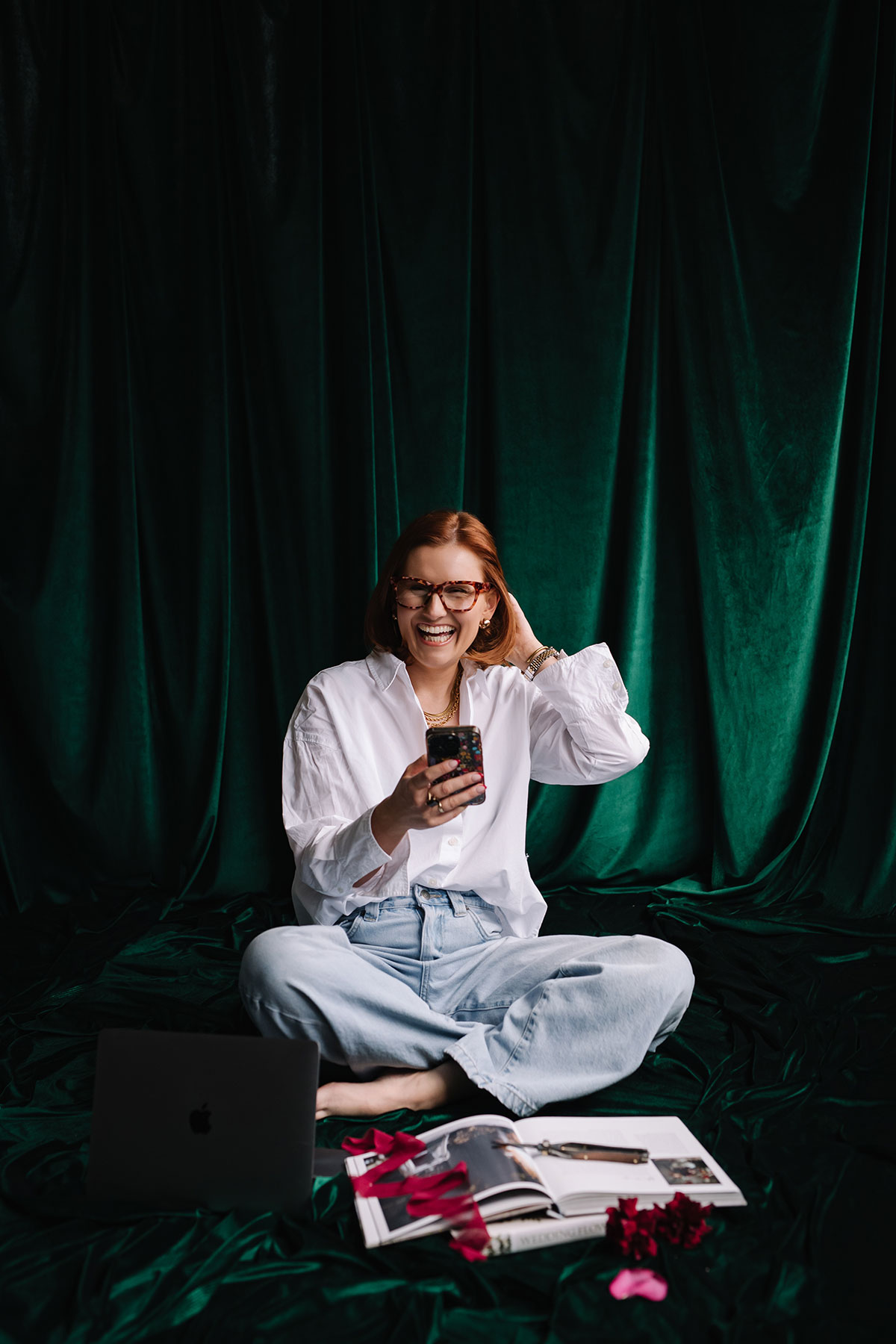 Woman with red hair and glasses sitting cross-legged in front of emerald green draped fabric, smiling while holding a phone, with magazines, ribbon and a laptop laid out beside her