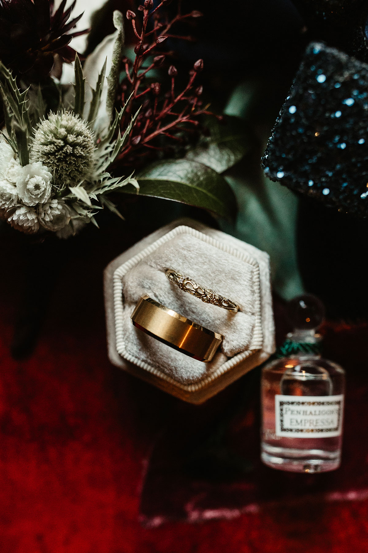 Close-up of gold wedding bands in a velvet ring box beside textured white and burgundy flowers and a perfume bottle