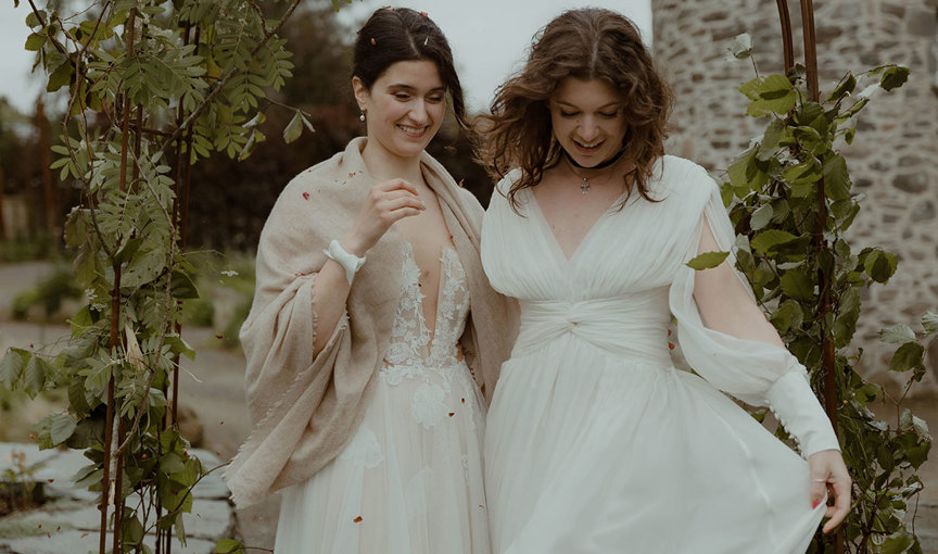 two brides standing closely side by side under an arch covered in green leafy foilage. There is an old stone castle wall behind them and a scattering of dark red petal confetti on their dresses