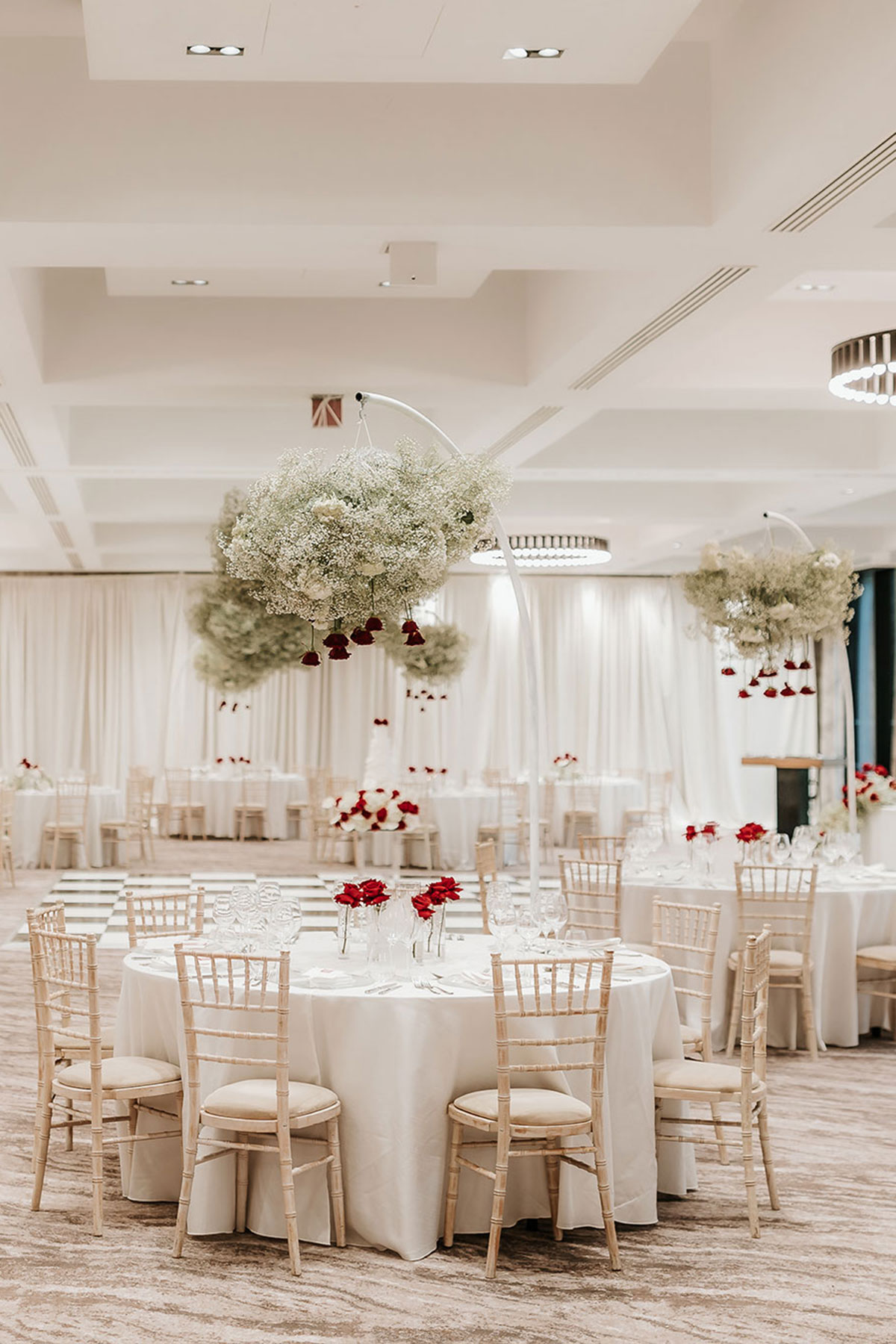 a selection of white tablecloth round tables set in a large room and decorated with single stem roses as well as a hanging floral installation placed by each individual table