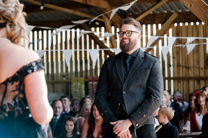 A Man In A Black Kilt Outfit Standing In A Wooden Slatted Building Looking At A Bride Wearing A Black Wedding Dress During Their Wedding Ceremony