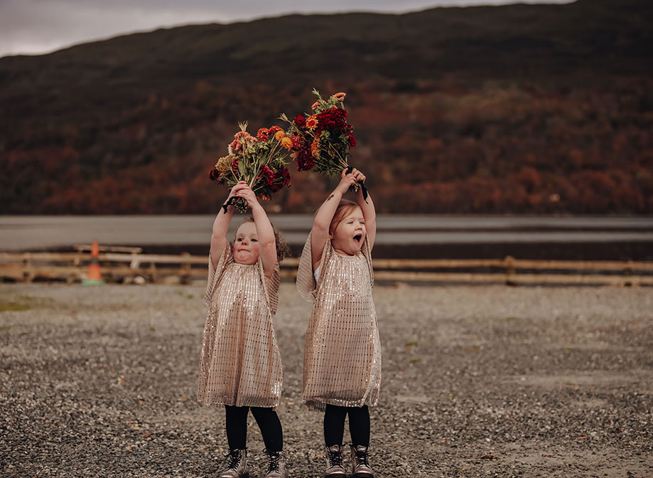 two young girls wearing gold sparkly dresses raising bunches of flowers above their heads