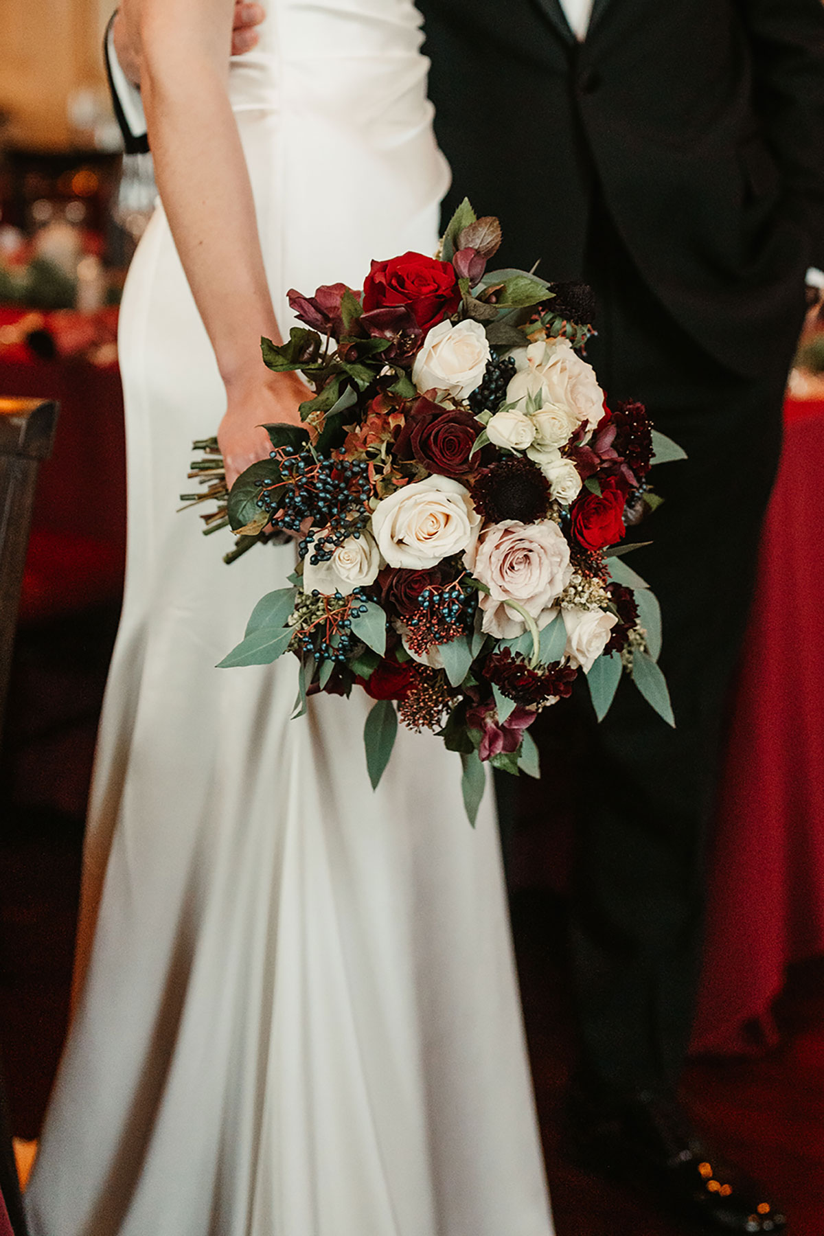 Bridal bouquet of red, blush and ivory roses with winter berries and greenery, held against a satin gown.
