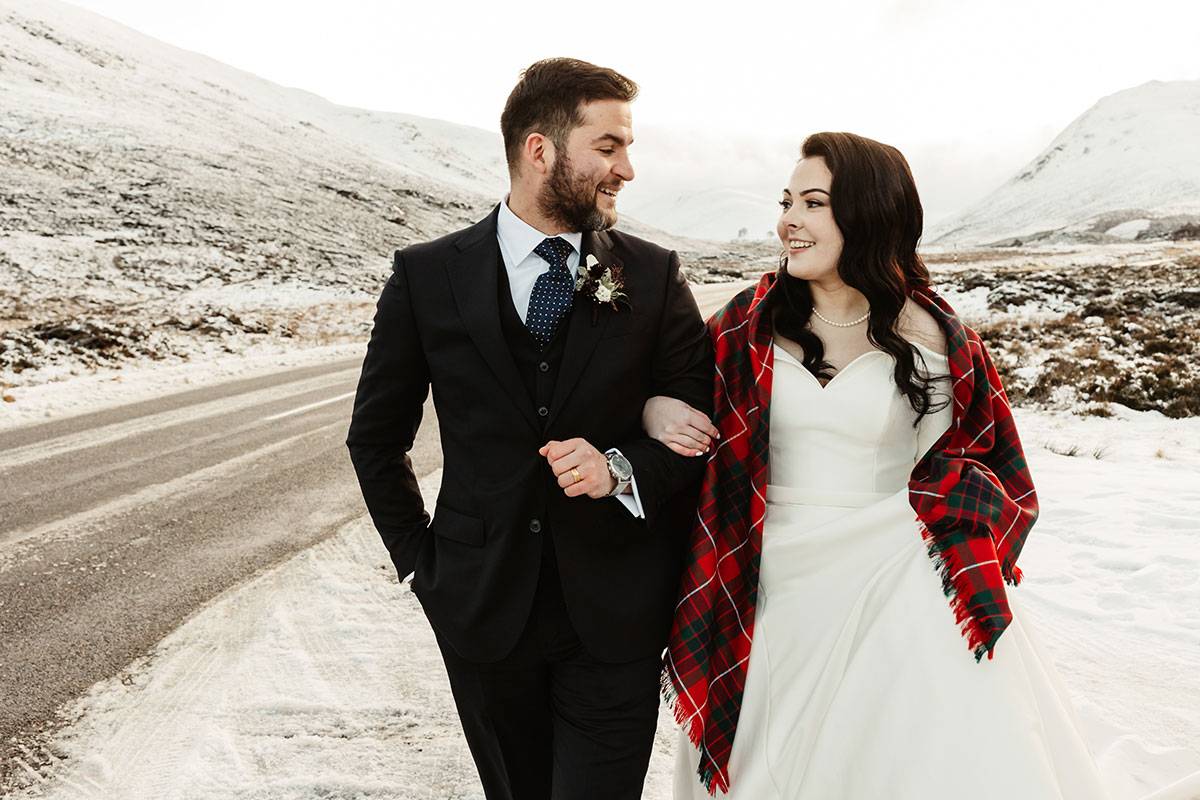 Bride and groom walking side by side on a wintry road, the bride wearing a red tartan shawl and smiling at the groom