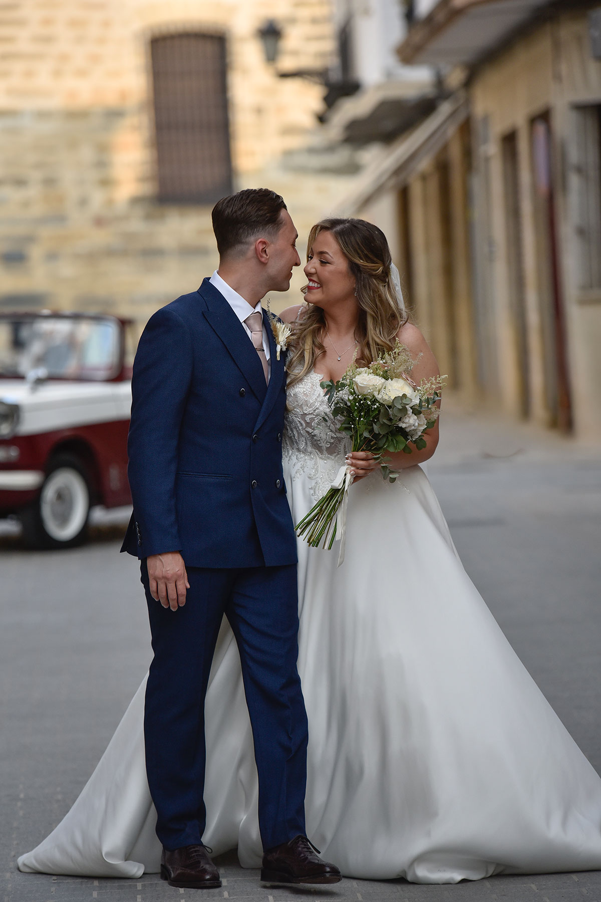 groom in blue suit and bride in ballgown with bouquet of white flowers stare into each other's eyes and smile