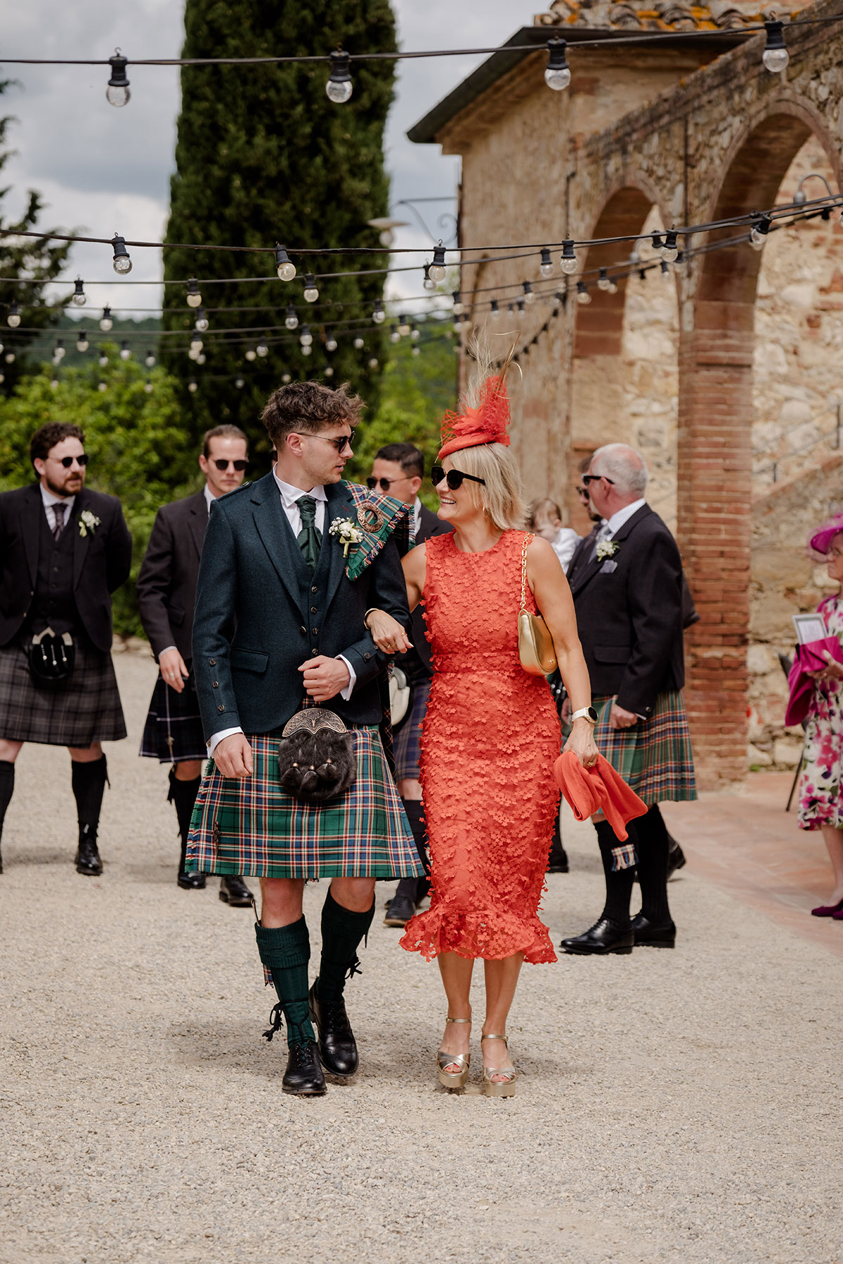 groom in blue, green and red tartan kilt outfit walks arm in arm with mum in orange midi dress and matching orange percher hat