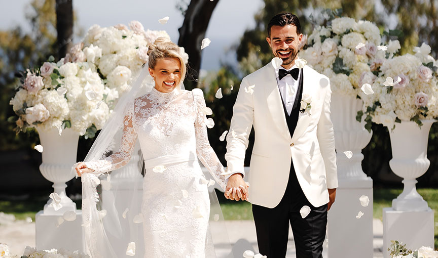 A bride and groom walking hand in hand through white confetti amid white urns filled with flowers.