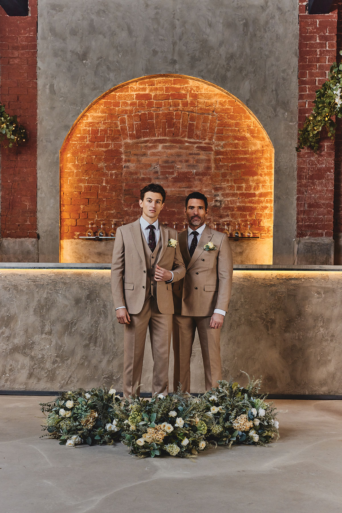 two men stand in tan coloured suit against stone backdrop and in front of floral display