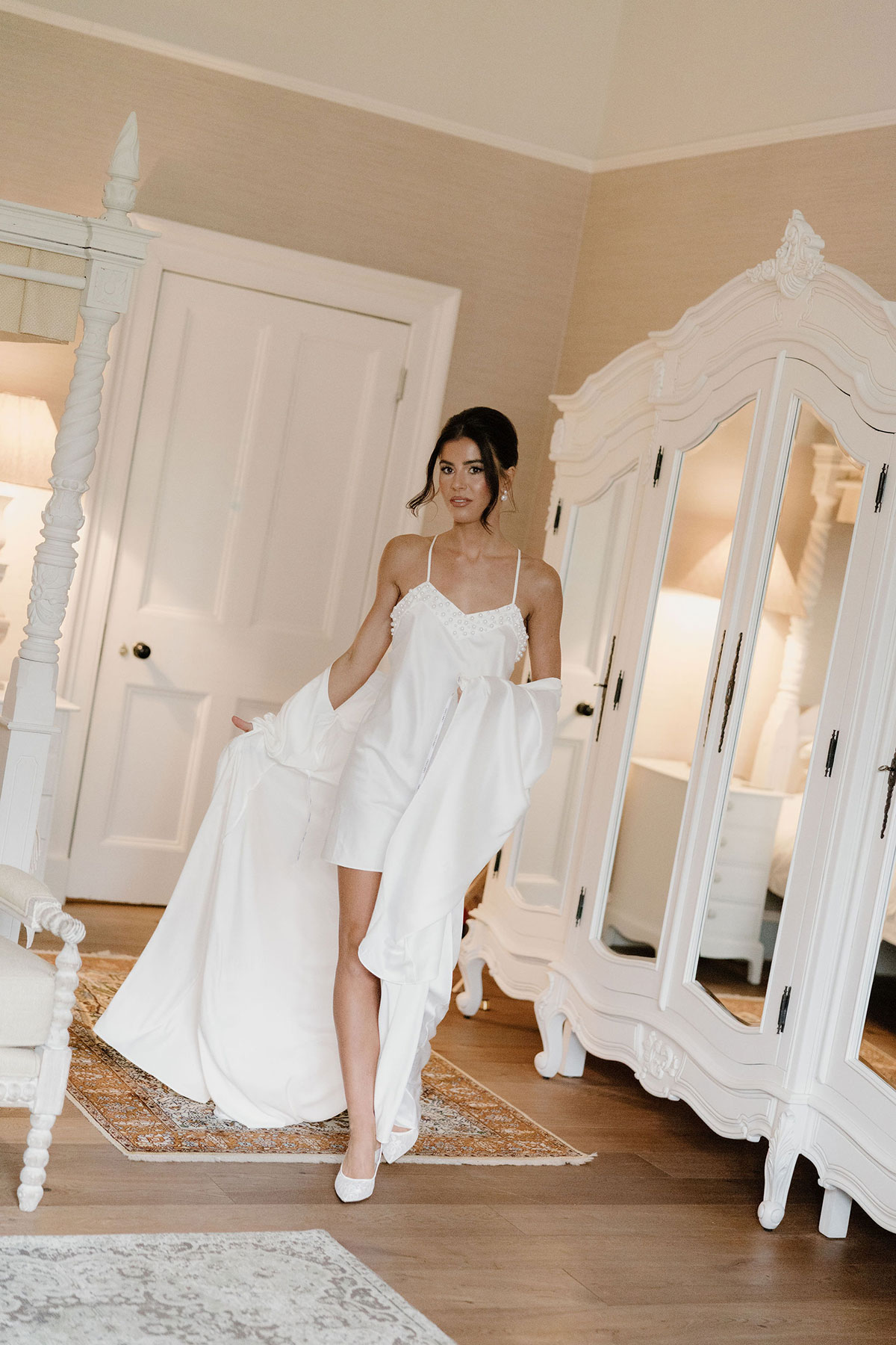 Bride wearing a satin robe and short slip dress standing in a bedroom beside a mirrored wardrobe