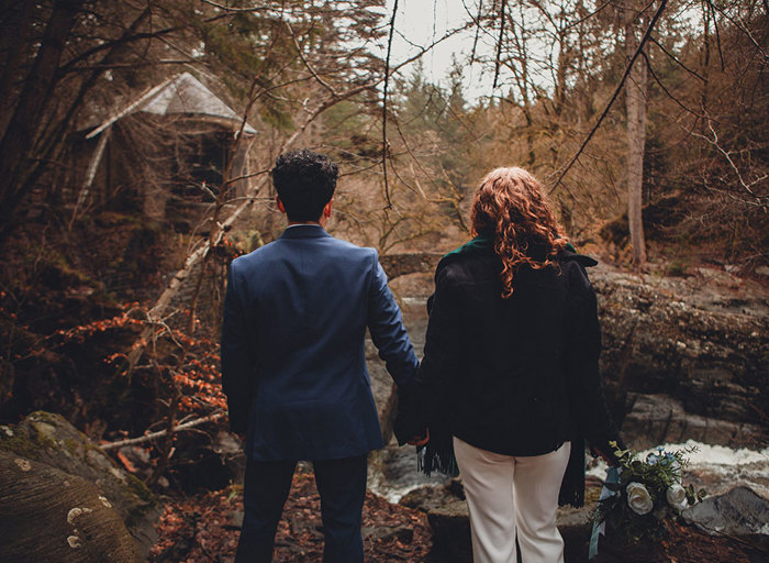 A couple facing away from the camera holding hands and looking out to a forest