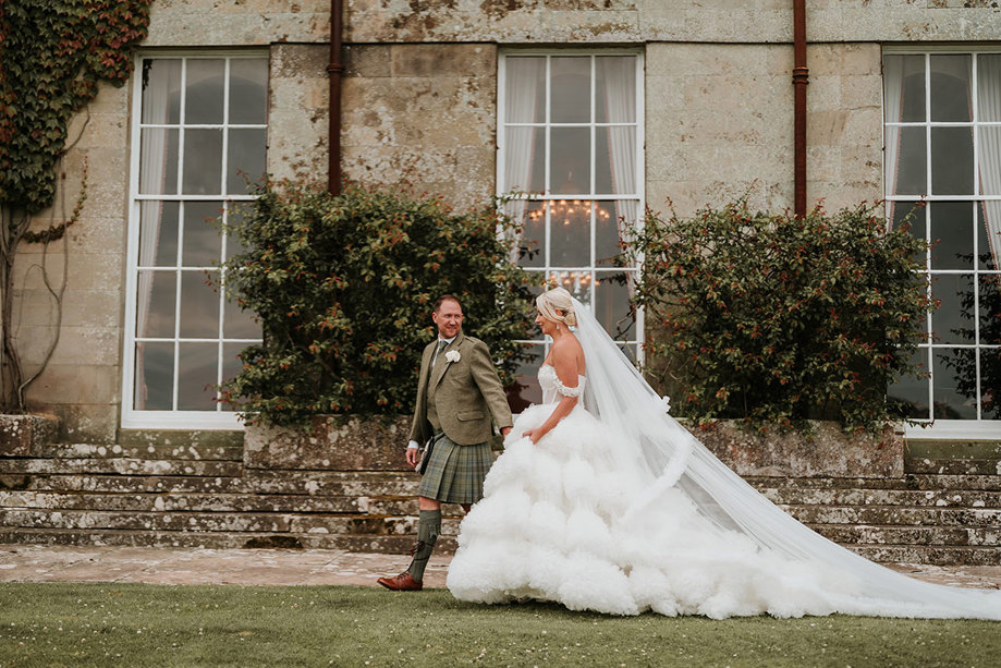 A bride wearing a wedding dress with a voluminous tulle skirt standing next to a groom wearing ga green kilt walking hand in hand in front of a building