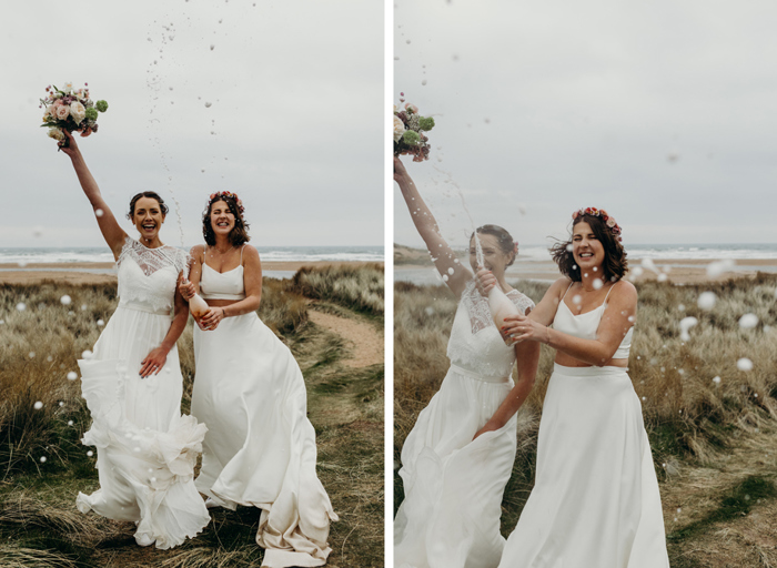 two elated brides standing on sand and scrubby-looking grass with sea in background performing a spray with a bottle of pink sparkling wine. Bride on left holds arm with bouquet aloft and bride on right wears flower crown and sprays the bottle