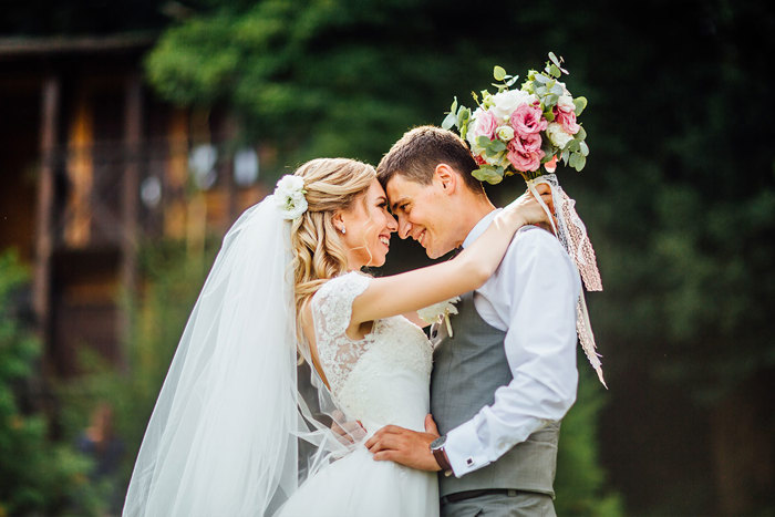 A bride and groom stand with their arms around each other and the bride is holding a white and pink bouquet