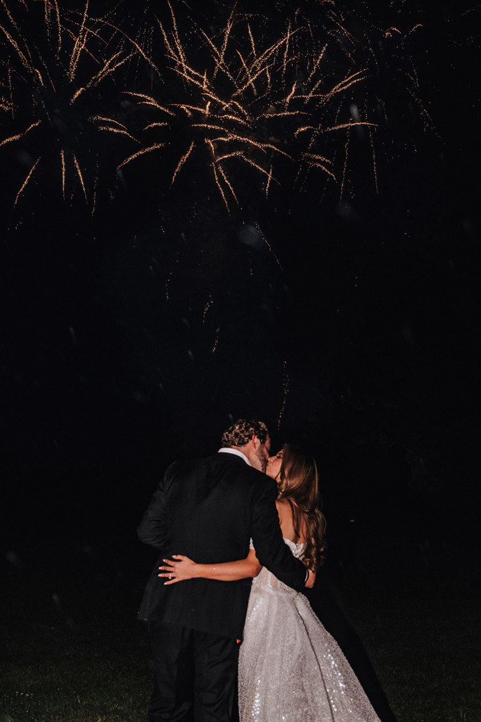 a bride and groom kissing as fireworks explode in the night sky.