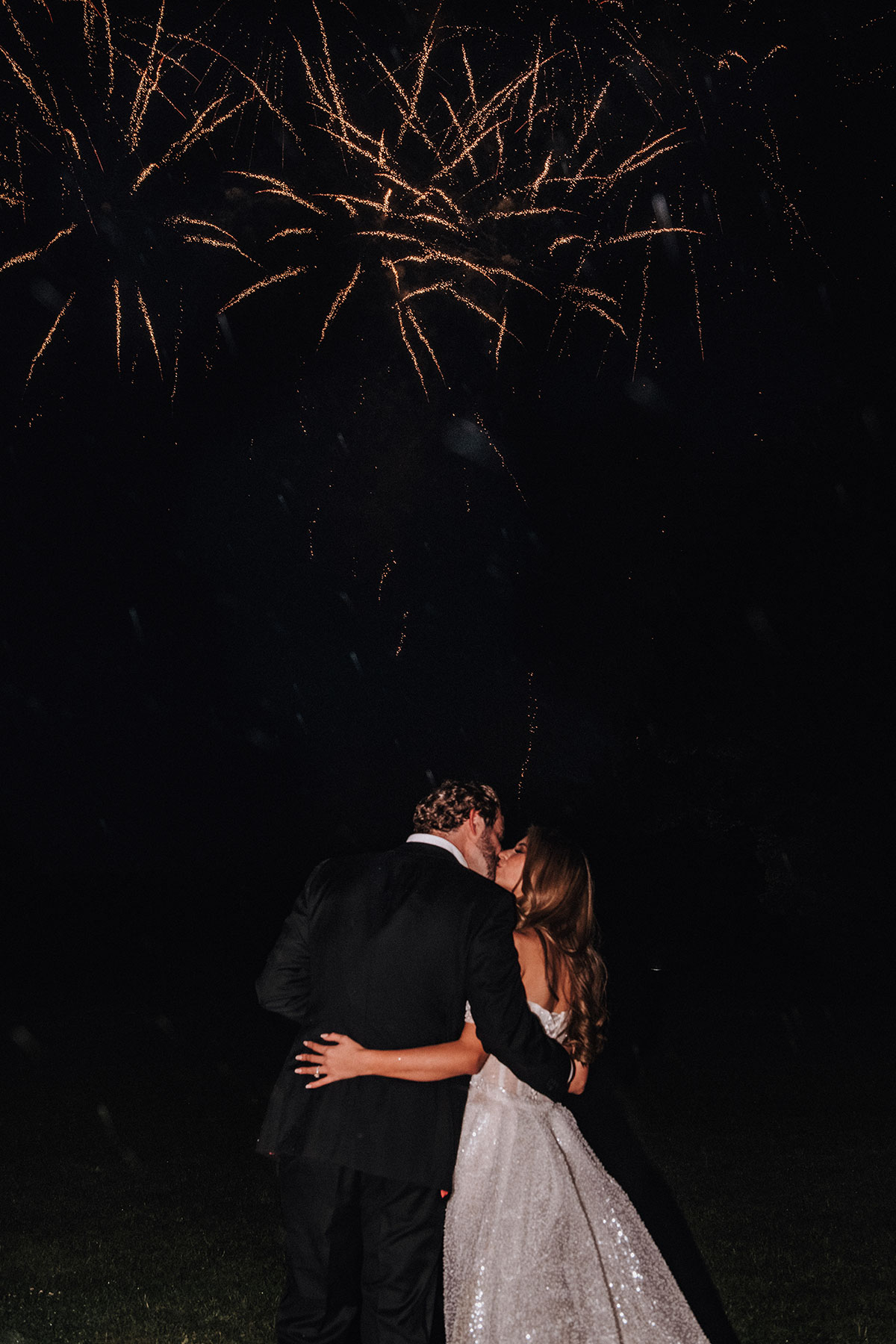 a bride and groom kissing as fireworks explode in the night sky.