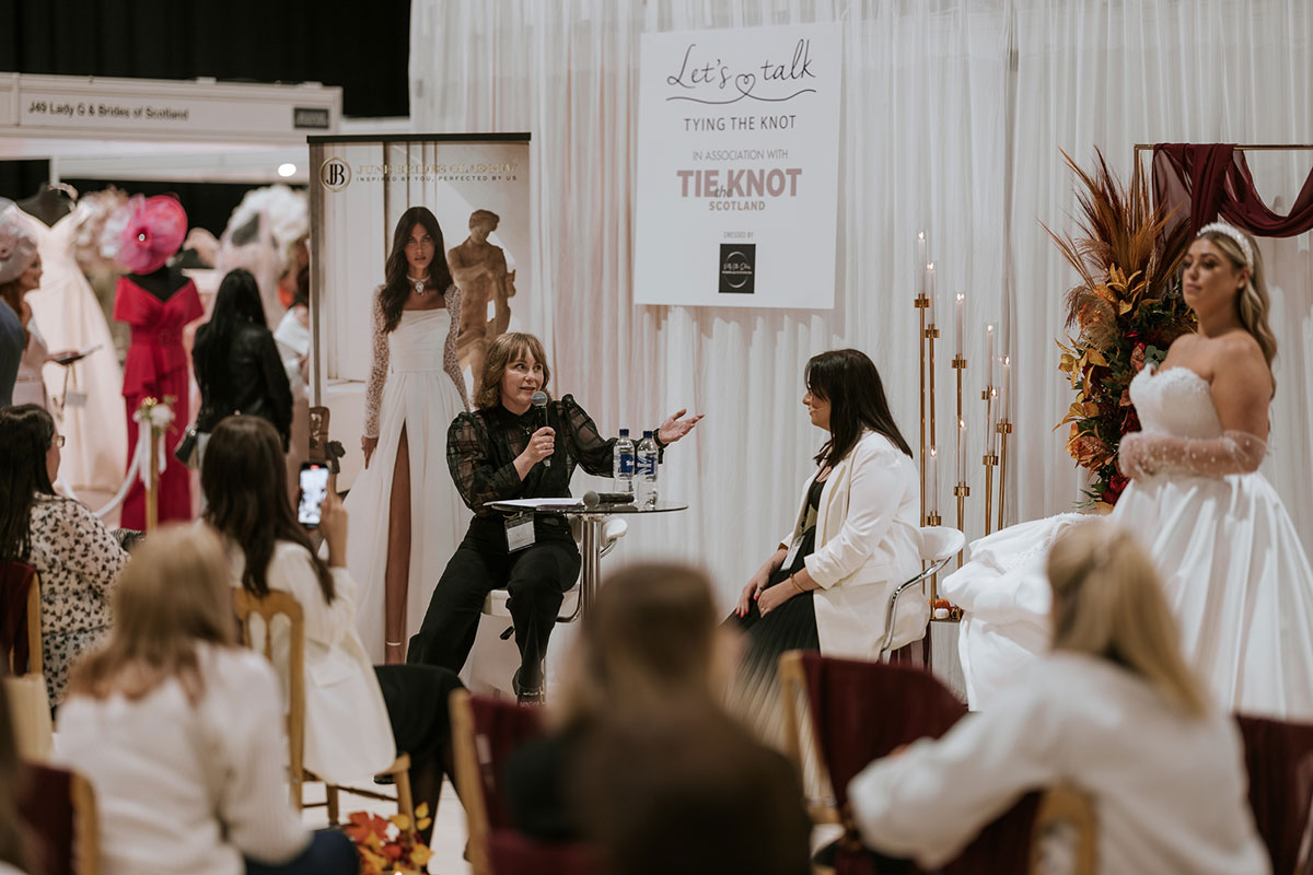 A person speaking to a person sitting at a table at The Scottish Wedding Show.