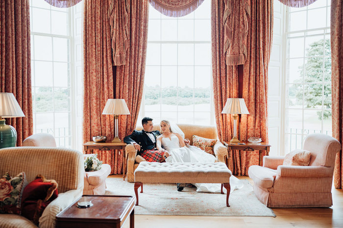 A bride in a wedding dress and a groom in a red kilt sitting on a couch in a room with curtains.