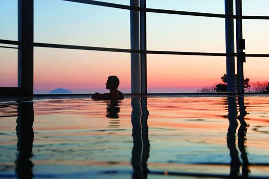 A man in a swimming pool looking out of large windows at a colourful sunset over a hill in the distance