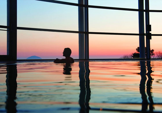 A man in a swimming pool looking out of large windows at a colourful sunset over a hill in the distance