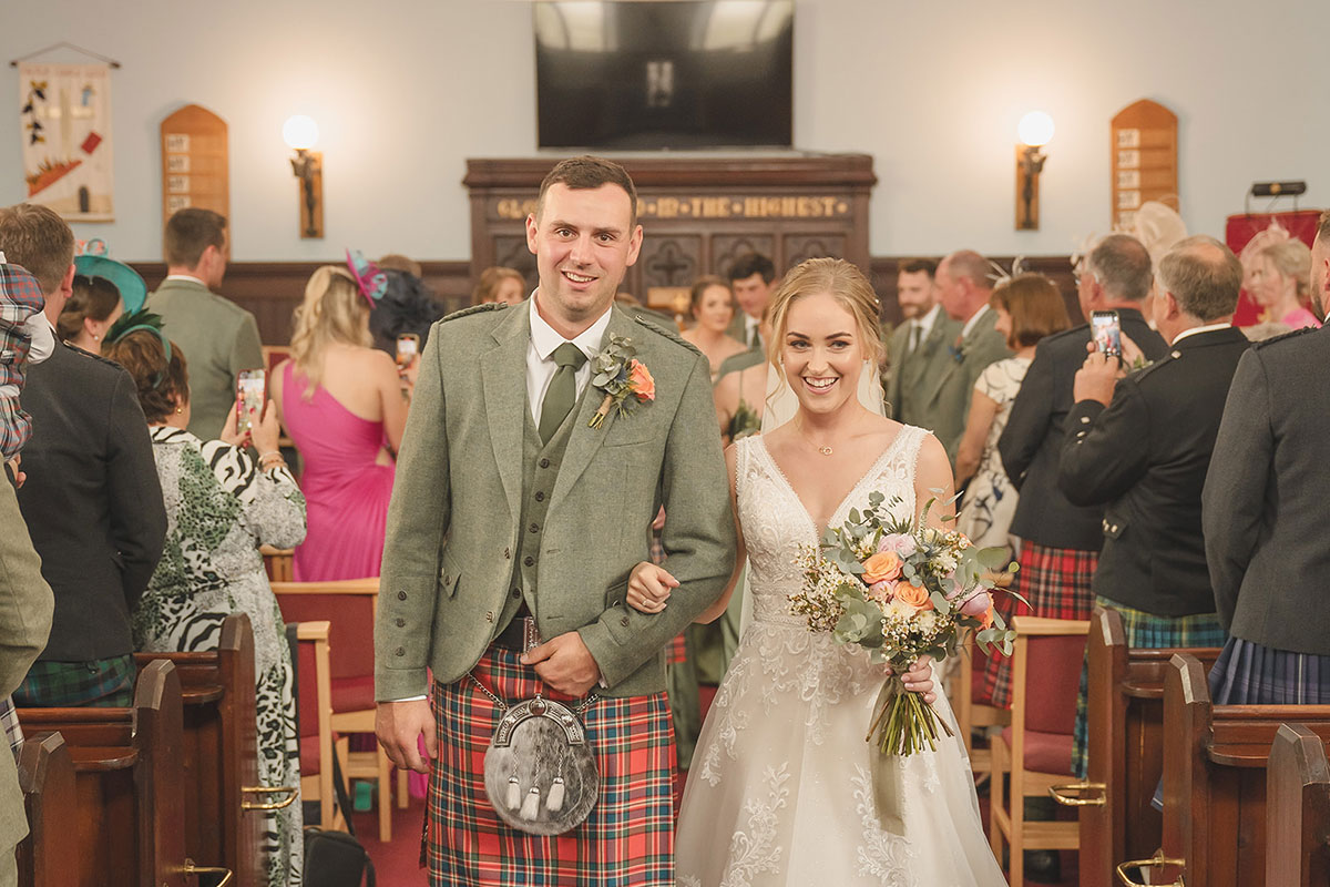 A man in a kilt and a bride in a wedding dress link arms and walk down the aisle