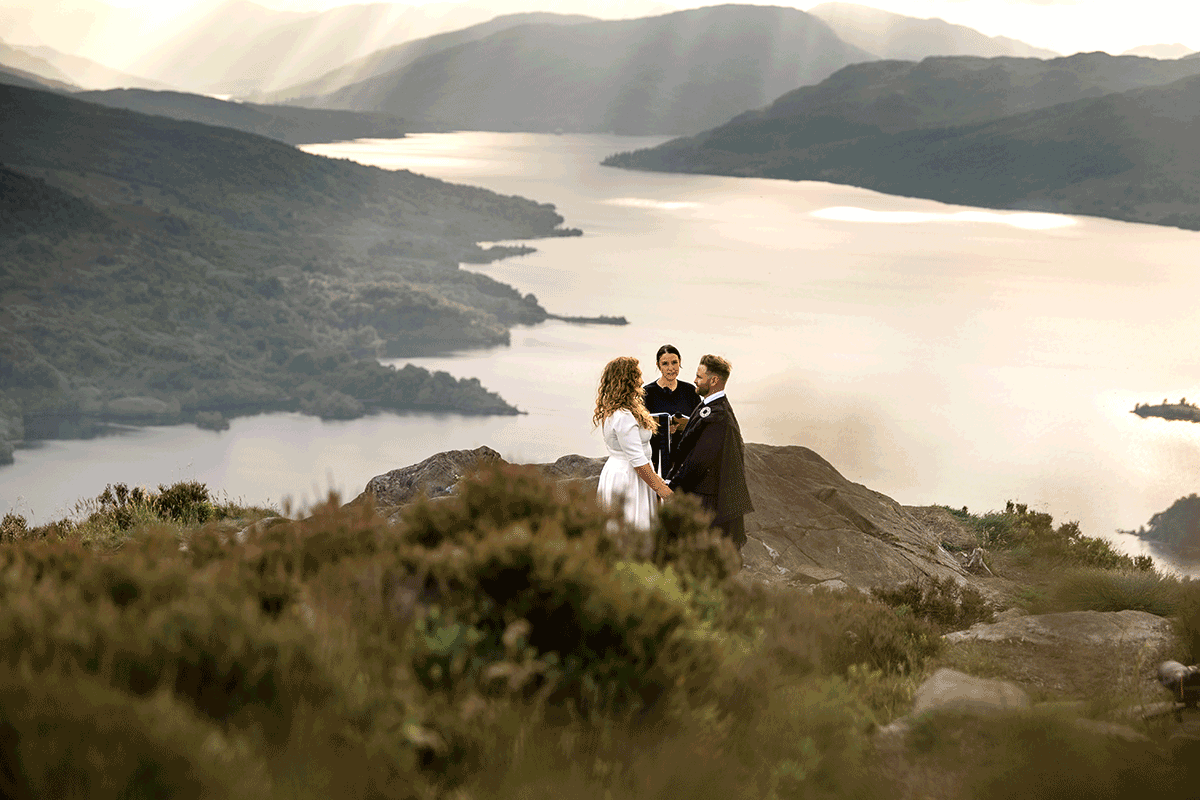 Bride, groom and celebrant stand on a rocky viewpoint above a Scottish loch, surrounded by dramatic Highland scenery and evening light