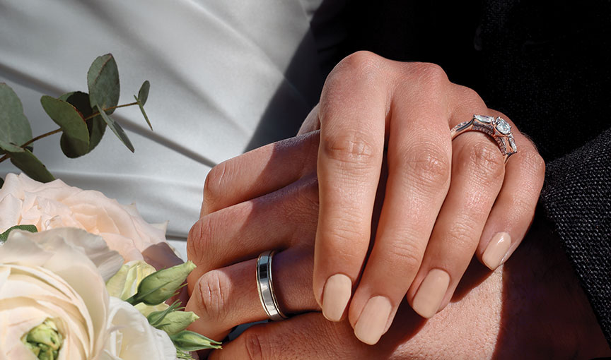 Close-up of newlyweds’ hands showing engagement ring, wedding bands and soft ivory florals.