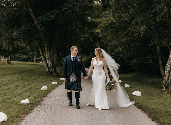 A groom in a dark coloured kilt and a bride in a fitted white dress with a long veil walk outside holding hands