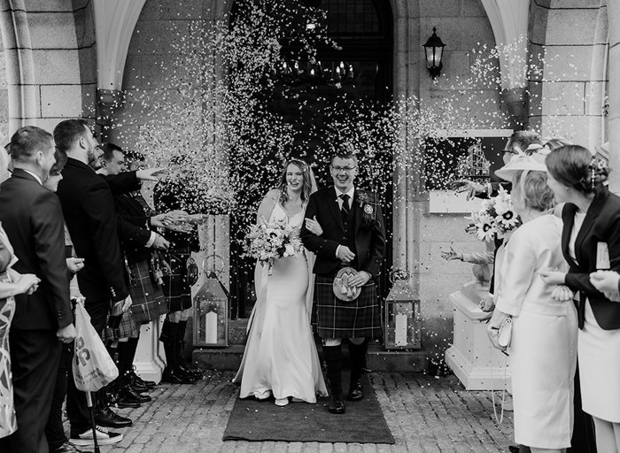 A black and white photo of a bride and groom walking through a door outside and being showered in confetti by their guests