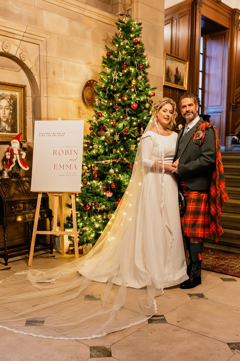 bride in white wedding dress and veil and groom in red and green tartan kilt pose closely side by side in front of green christmas tree with red decorations and a white welcome sign with their names on it