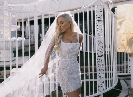 bride in embellished white mini dress poses in front of white gates with her long veil cast out to one side