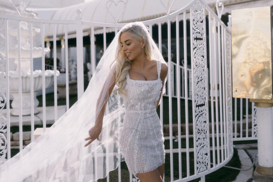 bride in embellished white mini dress poses in front of white gates with her long veil cast out to one side