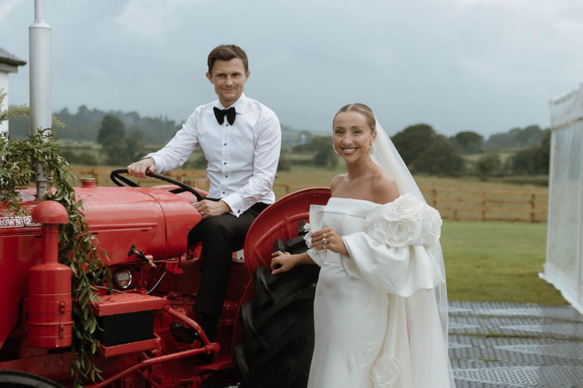 Bride and groom posing with a red vintage tractor at a countryside marquee wedding.