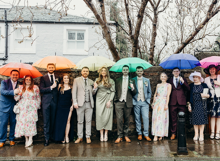 a group of people line up for a wedding photo along a wall under colourful umbrellas. There are trees and a building in the background