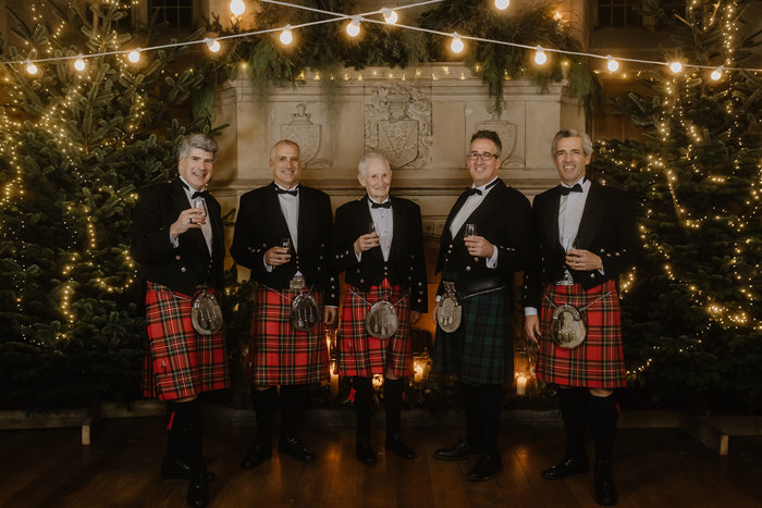 a row of men in red kilts standing in front of a large stone fireplace that's illuminated by fairy lights and has Christmas trees either side.