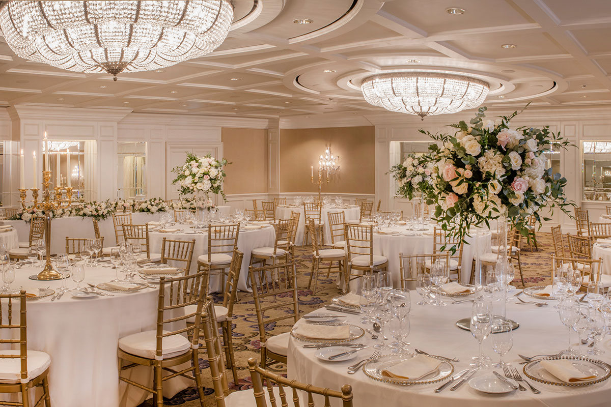 gold patterned carpet room is filled with round white tablecloth tables, gold chairs with white cushions and heightened centrepieces with pastel flowers and large ceiling chandeliers