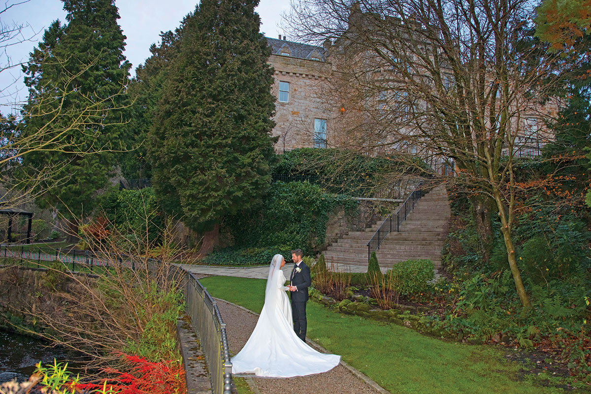 A bride and groom standing on a garden path surrounded by mature trees and landscaped grounds.