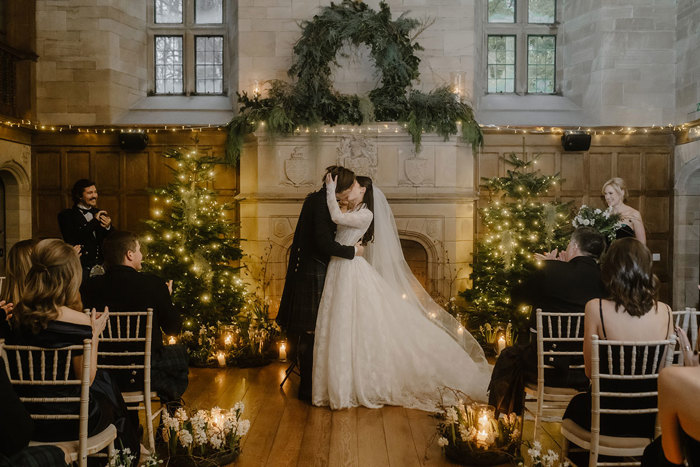 a kissing bride and groom standing in front of a large stone fireplace that's illuminated by fairy lights and has Christmas trees either side.