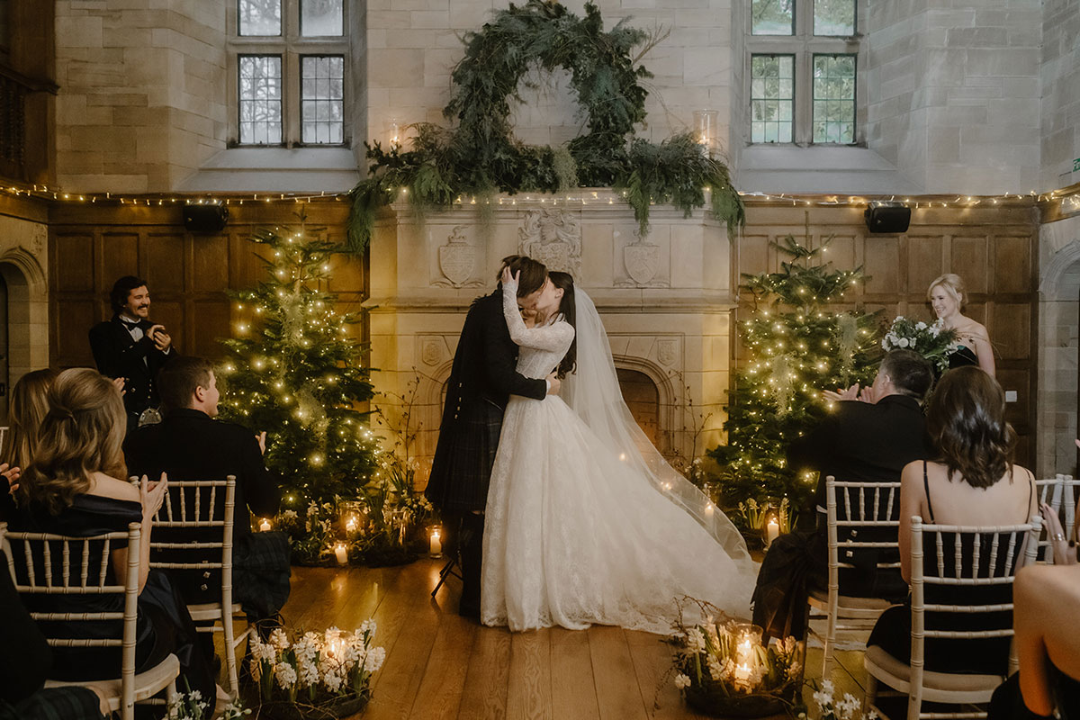a kissing bride and groom standing in front of a large stone fireplace that's illuminated by fairy lights and has Christmas trees either side.