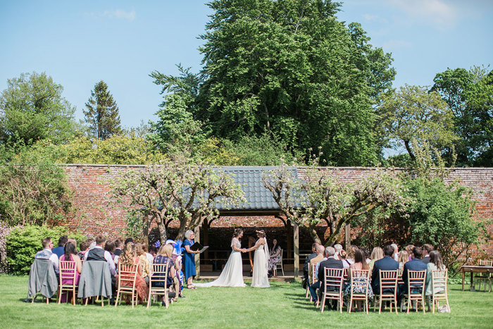 two brides and rows of seated wedding guests during an outdoor wedding ceremony at Byre at Inchyra. There is a large red brick wall and trees in the background.