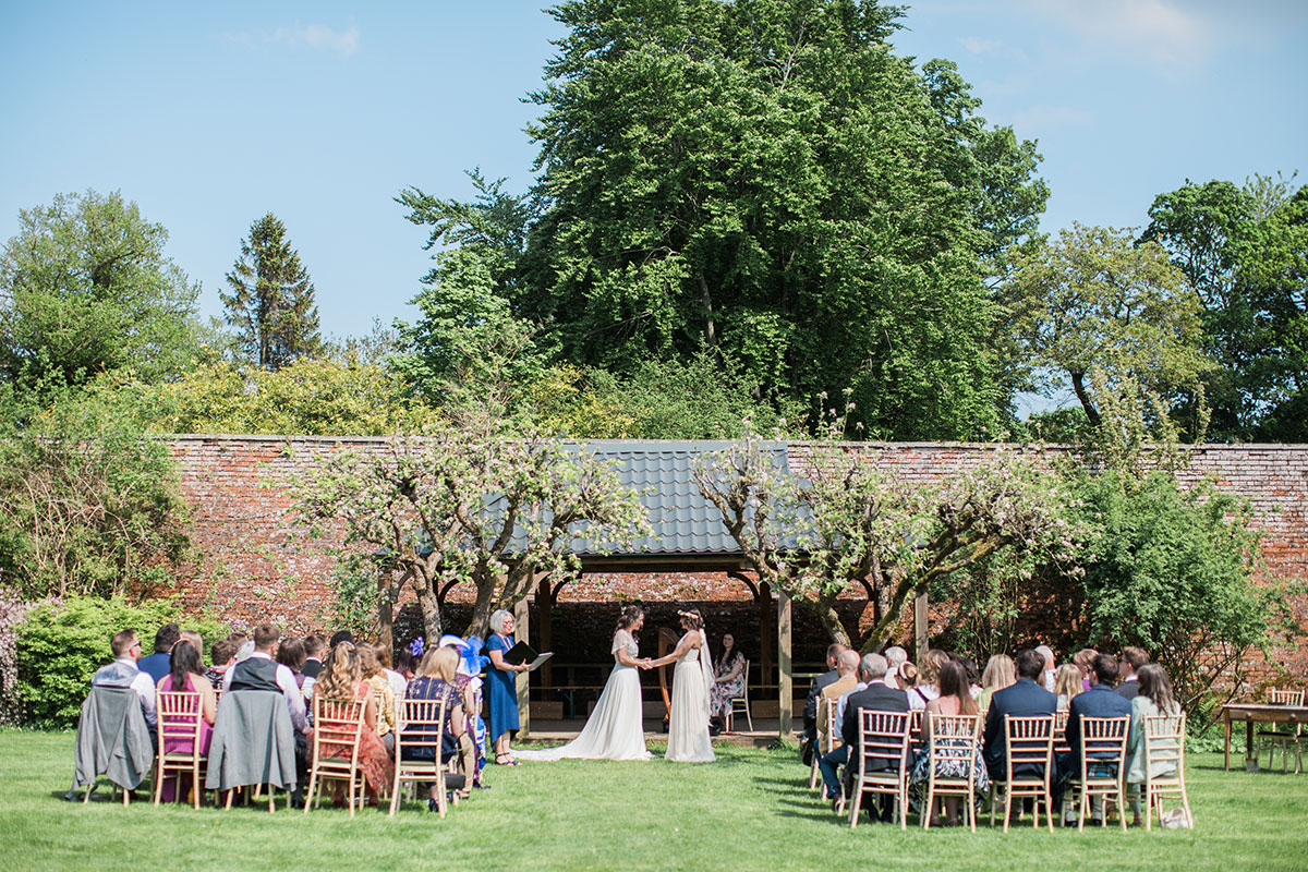 two brides and rows of seated wedding guests during an outdoor wedding ceremony at Byre at Inchyra. There is a large red brick wall and trees in the background.