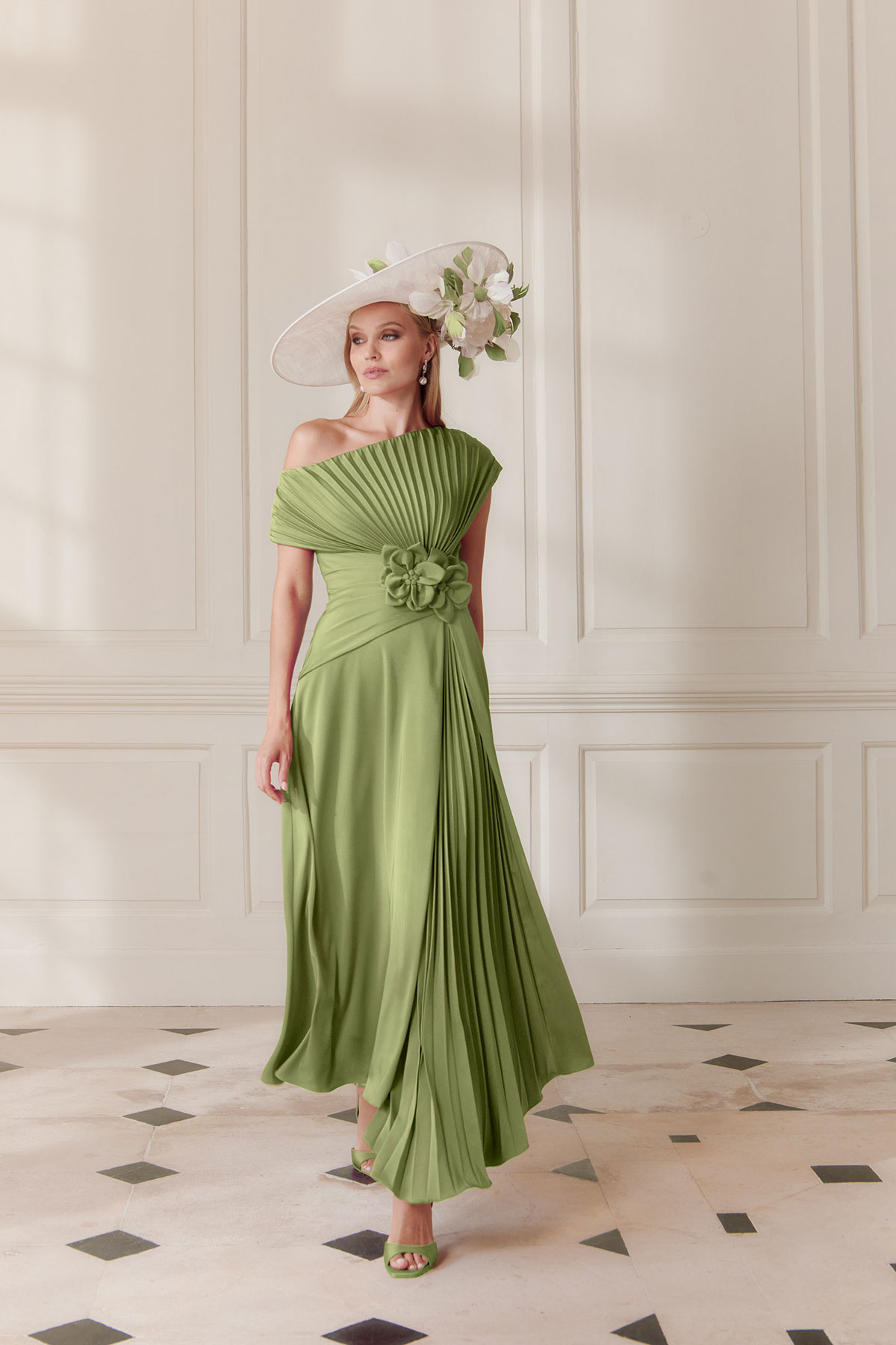 Model wearing a fern green off-the-shoulder pleated midi dress with floral waist detail and wide-brim ivory hat, standing against a neutral panelled wall