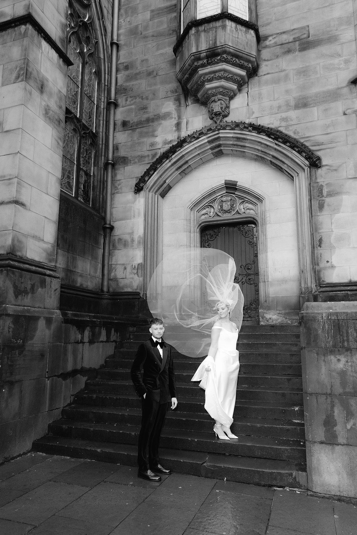 Bride and groom standing on stone steps outside a historic Edinburgh building, with the bride’s veil dramatically billowing in the wind