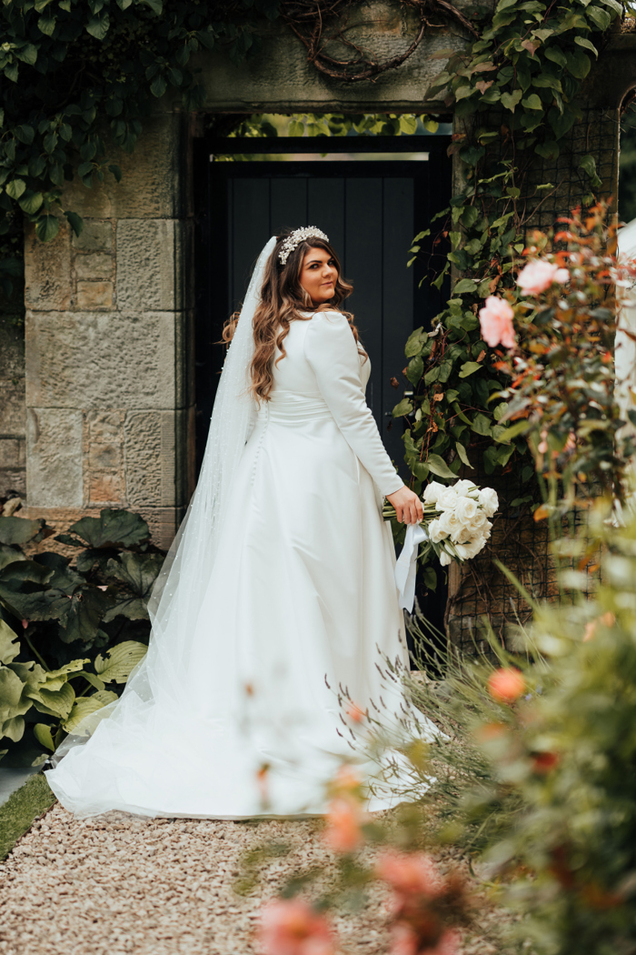 A bride wearing a long sleeve dress, sparkly hairband and veil faces away from the camera and looks back over her shoulder as she stands in a garden